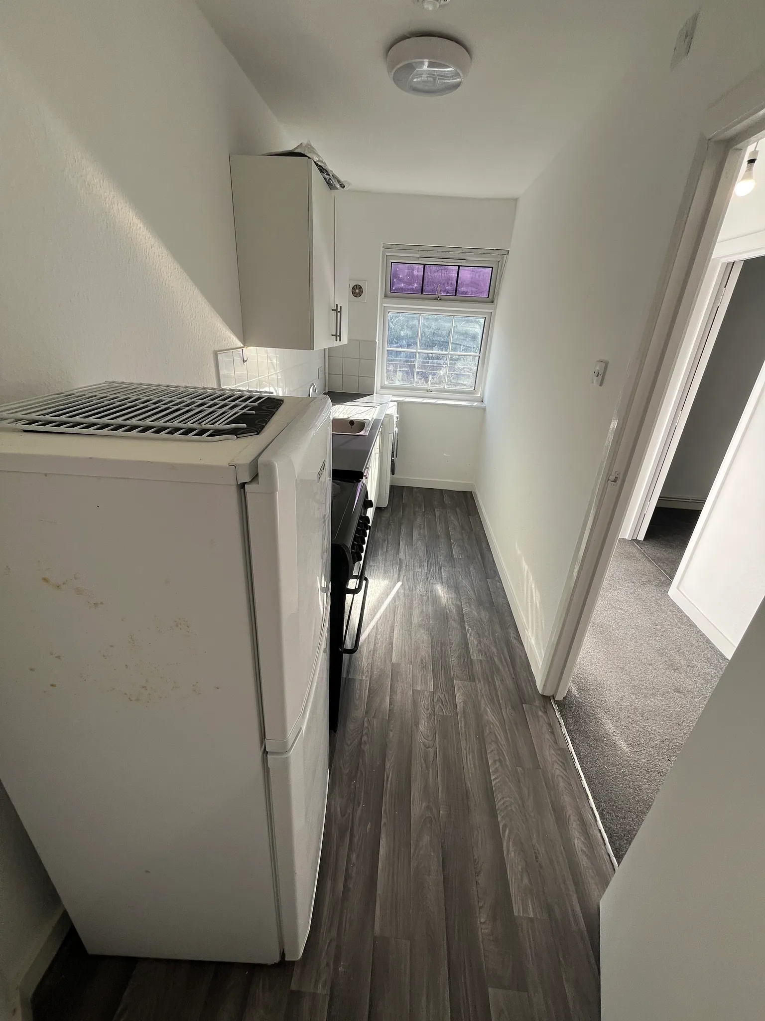 Narrow kitchen corridor with a refrigerator and stove on the left, wall cabinets, a small sink under a window, and wood-look flooring.
