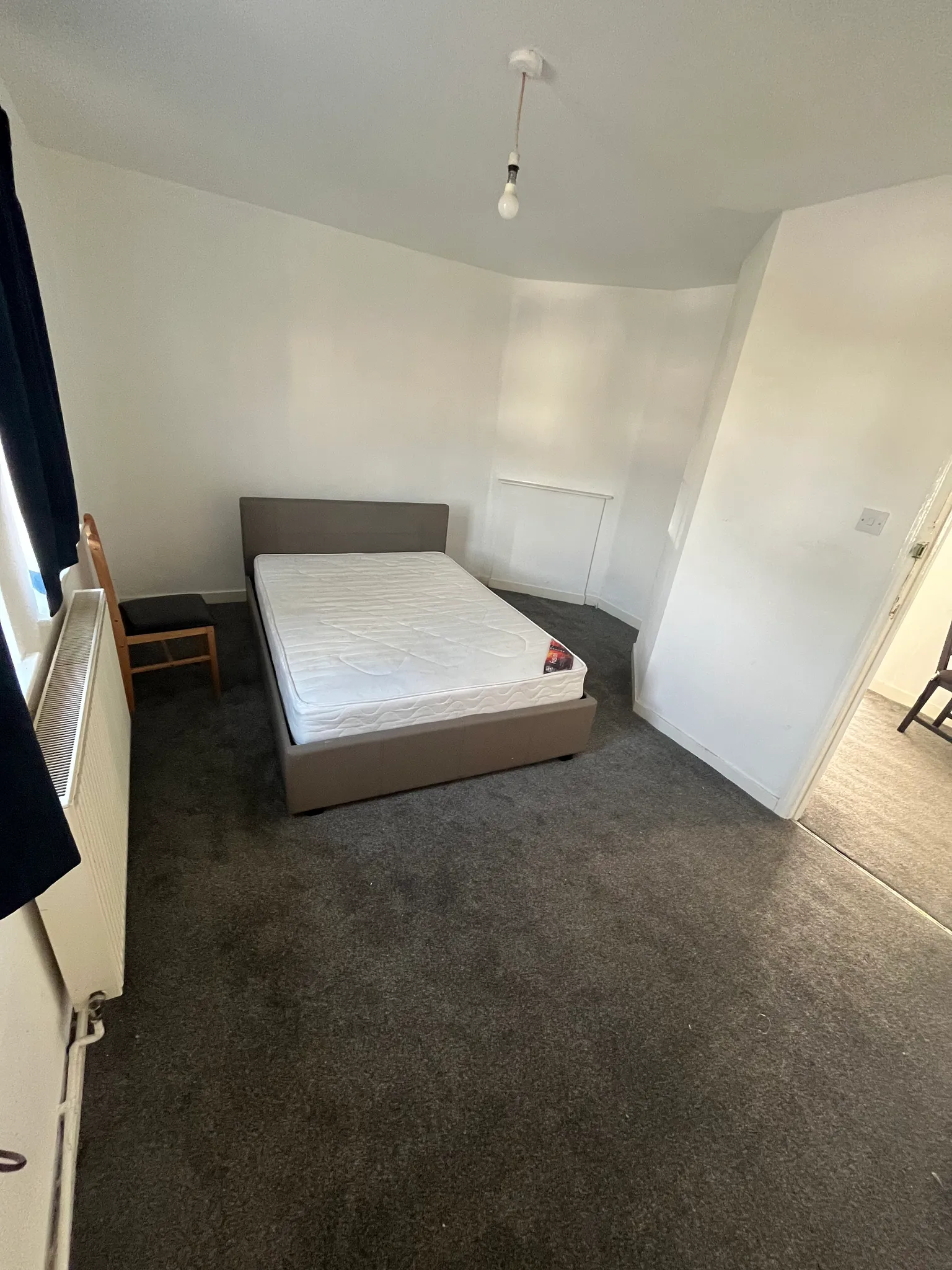 Bedroom with a double bed frame and mattress, a wooden chair by a window with radiator, grey carpet and white walls.
