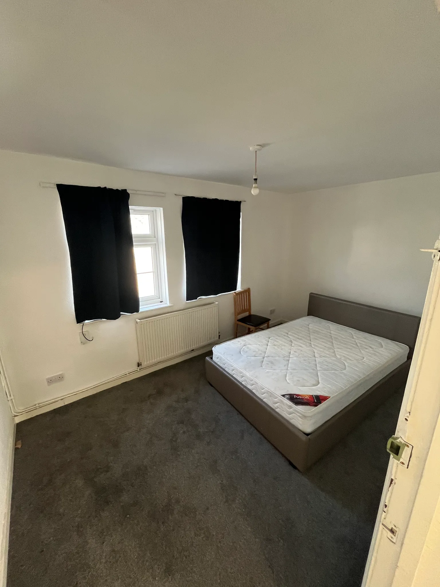 Bedroom with a double bed and mattress, two covered windows with dark curtains, a radiator, and a wooden chair.
