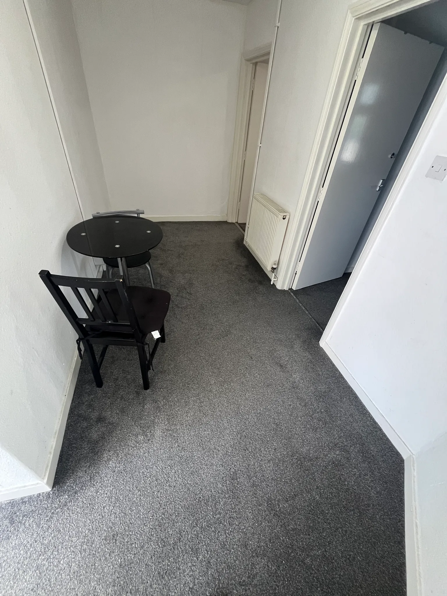 Narrow hallway with a small round table, one chair, a wall radiator and a partially open door.
