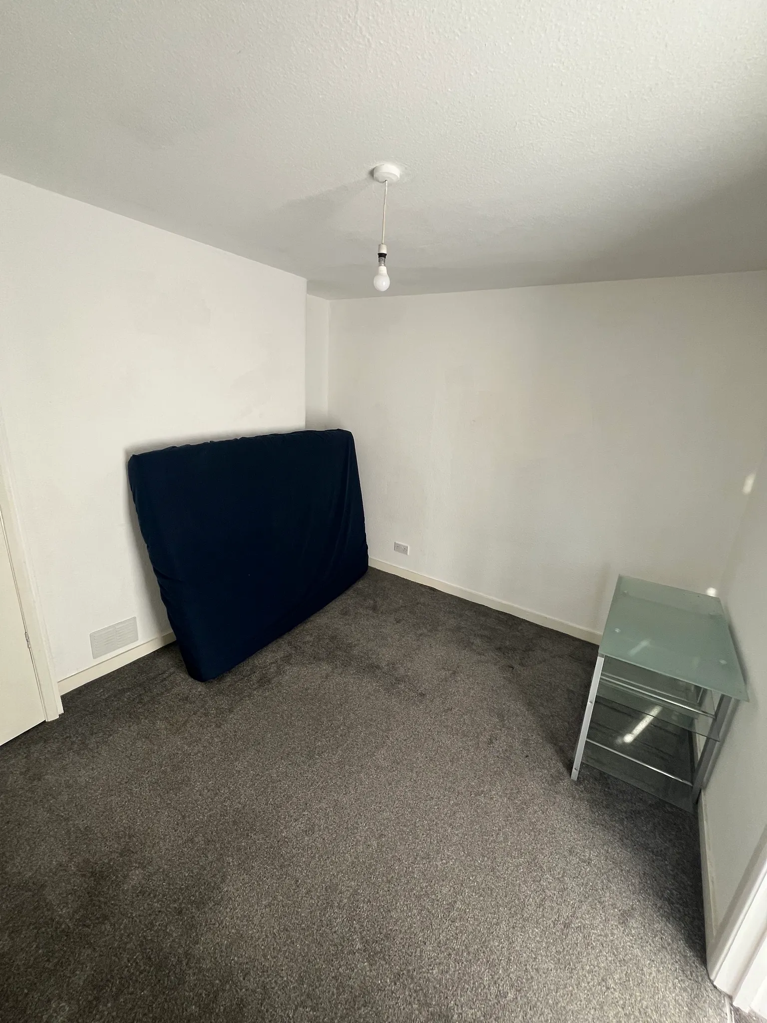 Small bedroom corner with a bare ceiling light bulb, a mattress propped against a white wall, grey carpeted floor and a small glass shelving unit